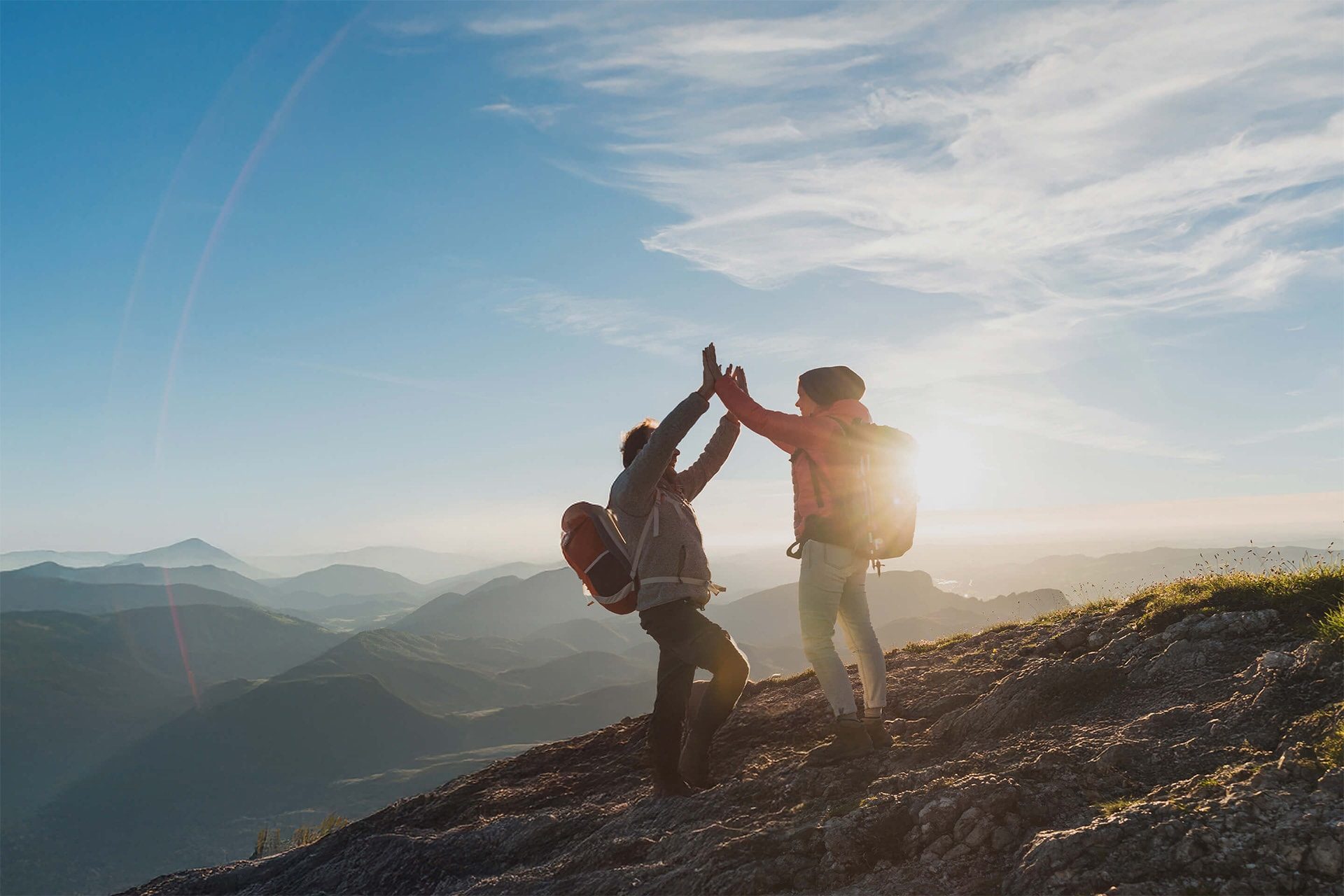 Zwei Wanderer, die einen Gipfel mit einem High-Five feiern, ihre Silhouetten zeichneten sich gegen die untergehende Sonne über einer bergigen Landschaft ab. 