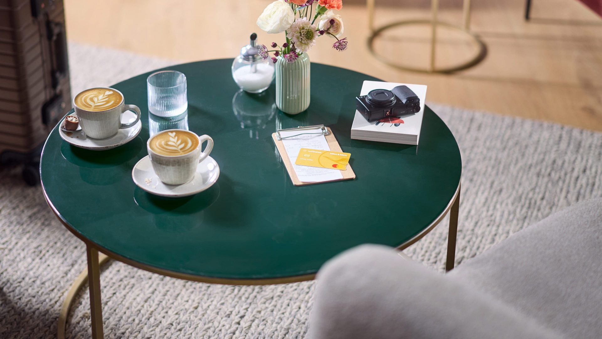 Die Miles & More Gold Credit Card liegt auf einem Kassenbon auf einem Lounge-Tisch. Daneben sind Fotoappart, Buch und Kaffeetasse. 