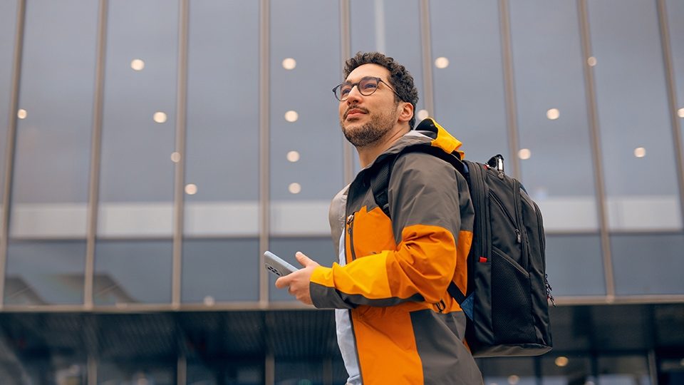 Person mit Rucksack und Smartphone vor einer modernen Glasfassade 