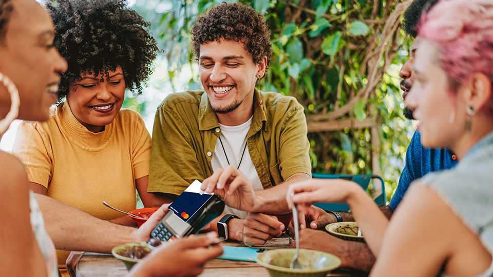 Gruppe sitzt am Tisch und bezahlt kontaktlos mit einer Miles & More MyFlex Credit Card. 